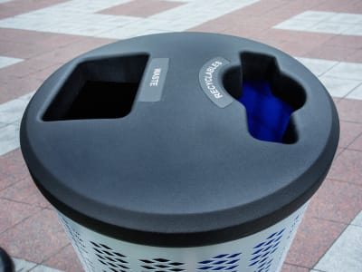 top angled view of a double stream waste and recycling container with a metal body and black plastic lid on a city sidewalk