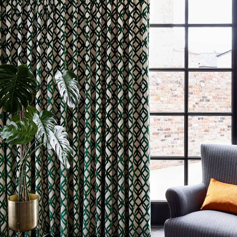 Heavy patterned curtains with green-and-black lattice on beige, drawn closed, filtering light; beside a mullioned window showing a brick wall in a living-room corner with monstera and gray armchair.