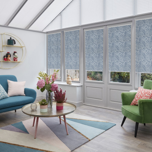 Patterned blue roller blinds covering tall conservatory windows, lowered to mid-height and filtering daylight; contemporary sunroom with teal sofa, green armchair, round coffee table, floral arrangements, and pastel rug.