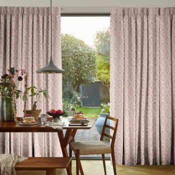 Pleated pink diamond-pattern curtains, drawn apart over a sliding glass door; filtering soft daylight into a dining room with wooden table, pendant lamp and a sunlit garden view.