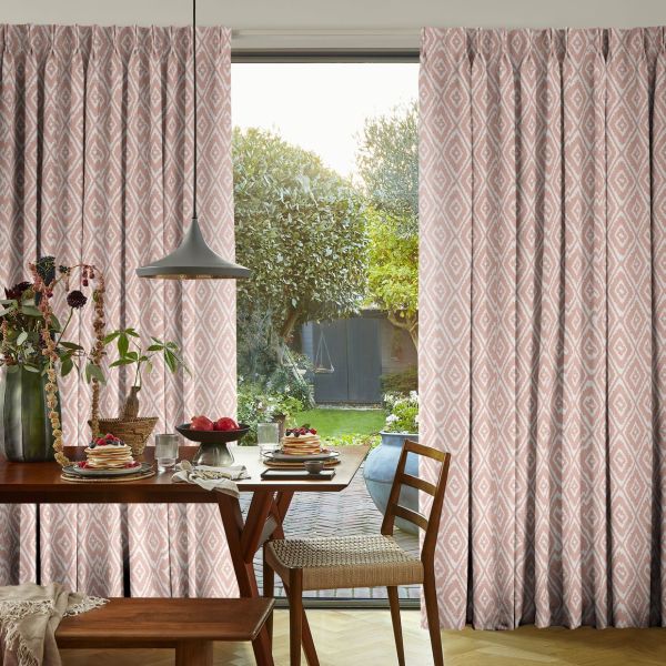 Pleated pink diamond-pattern curtains, drawn apart over a sliding glass door; filtering soft daylight into a dining room with wooden table, pendant lamp and a sunlit garden view.