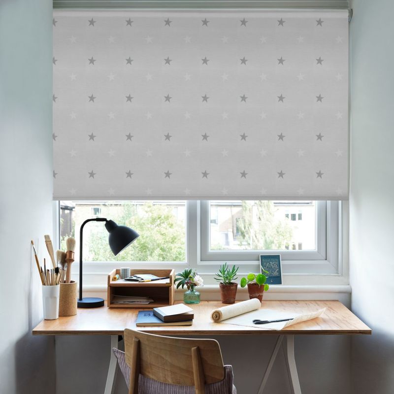 Pale grey roller blind with star pattern, lowered and softly filtering daylight; wooden desk beneath with lamp, paintbrushes and small potted plants, trees and houses visible outside.