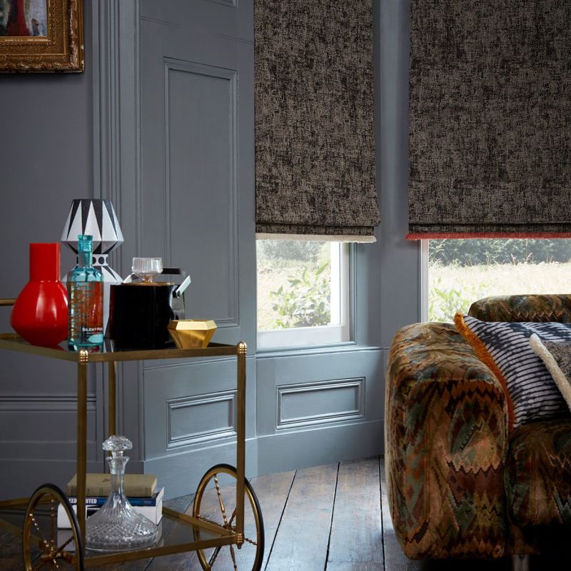 Textured dark grey Roman blinds, lowered over two tall windows with red trim, partially blocking light; in a living room with grey paneled walls, patterned sofa, wooden floor, garden visible below.
