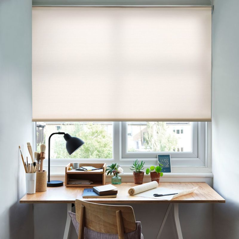 A beige roller blind lowered nearly fully over a double window, textured; filtering soft daylight into a small home workspace with wooden desk, lamp, art supplies and potted plants.