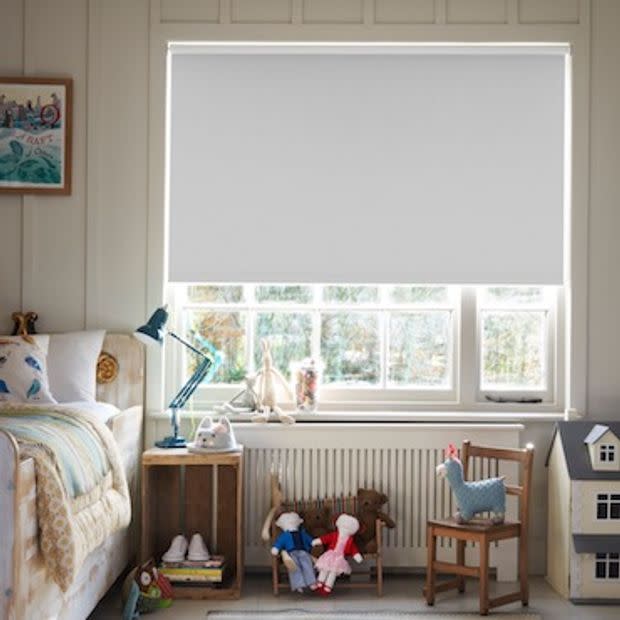 Roller blind (light grey, opaque) pulled down across upper window, diffusing daylight while lower panes remain visible. Child’s bedroom with bed, lamp, toys and small garden view beyond.