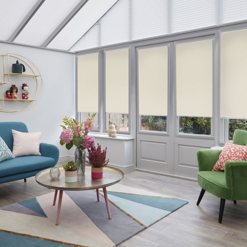 Beige fabric roller blinds lowered two-thirds over tall glass doors and windows, with white pleated blinds in the pitched roof, filtering soft daylight into a colorful conservatory-style living room.