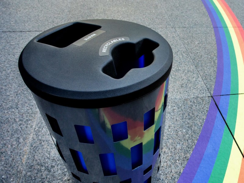 top angle view of a double waste and recycling bin with a metal body and black plastic lid sitting on a city sidewalk with a rainbow painted on the ground