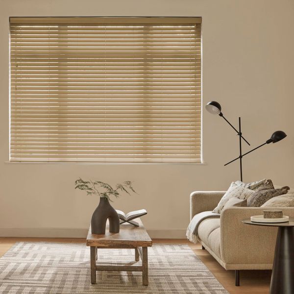 Horizontal beige Venetian blinds cover a large window, closed and gently filtering daylight; a neutral living room with a sofa, wooden coffee table, patterned rug and angled black floor lamp.