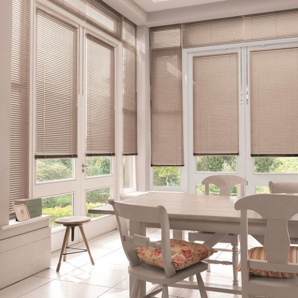 Beige horizontal blinds covering tall French doors and windows, lowered with slats mostly closed and softly filtering daylight; sunlit dining corner with wooden table, chairs, floral cushions and garden view.