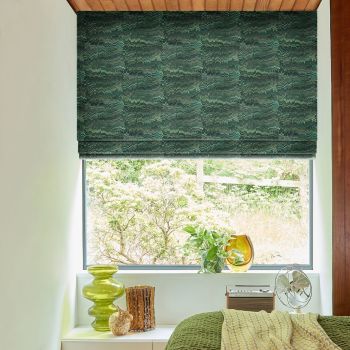 Green patterned Roman blind lowered in soft folds, partly blocking and filtering daylight, in a bedroom window over a sill with glass vases, a potted plant, small fan and green bed.
