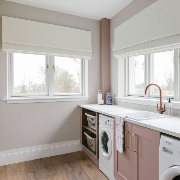 White Roman blinds, lowered and softly folded across two windows, filtering overcast daylight; laundry room countertop with pale‑pink cabinets, sink, washing machines, wooden floor and a view of bare trees.
Whirlpool