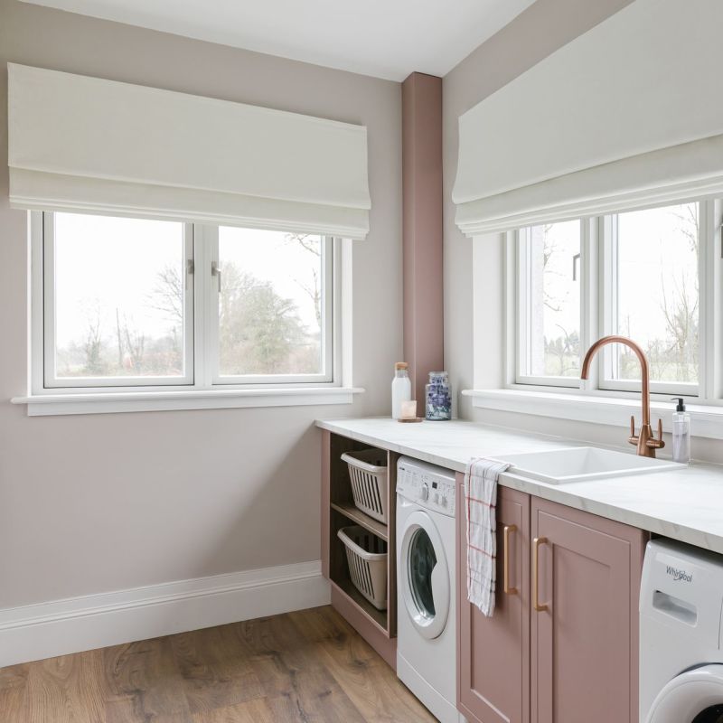 White Roman blinds, lowered and softly folded across two windows, filtering overcast daylight; laundry room countertop with paleâ€‘pink cabinets, sink, washing machines, wooden floor and a view of bare trees.

Whirlpool
