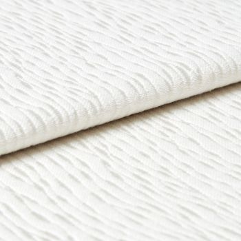 White wavy-ribbed fabric folded with an overlapping edge; fabric lies flat and layered. Close-up view under soft, diffuse light on a neutral surface, suggesting bedding or upholstery.