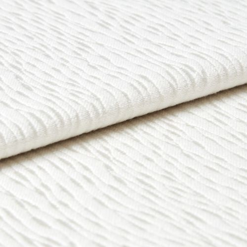 White wavy-ribbed fabric folded with an overlapping edge; fabric lies flat and layered. Close-up view under soft, diffuse light on a neutral surface, suggesting bedding or upholstery.