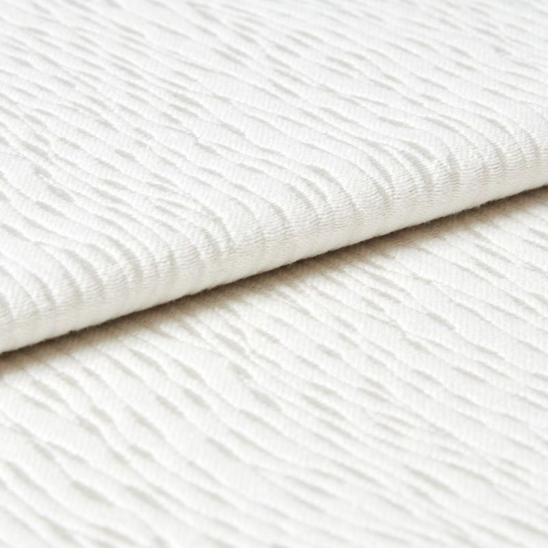 White wavy-ribbed fabric folded with an overlapping edge; fabric lies flat and layered. Close-up view under soft, diffuse light on a neutral surface, suggesting bedding or upholstery.