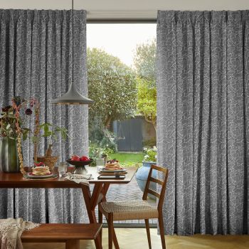 Patterned grey geometric curtains, pleated and drawn aside, framing and filtering sunlight; a wooden dining table set for breakfast with pendant lamp, opening onto a sunlit garden with trimmed trees.