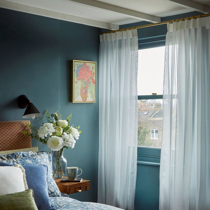 Sheer white curtains on a brass rod, drawn to frame a window and filtering daylight; bedroom with teal walls, patterned bedding, nightstand holding a vase of white flowers and a rooftop view.
