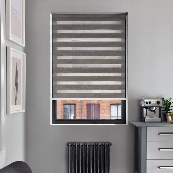 Gray zebra roller blind, lowered over window with alternating opaque and sheer bands, filtering daylight; framed artwork left, black radiator below, countertop with espresso machine to right in gray room.