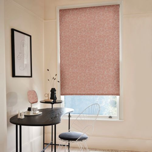 A pink roller blind patterned with white continuous-line faces, fully lowered over a tall window, softly filtering daylight in a minimalist desk/vanity corner with a black desk, wire chair and framed art.