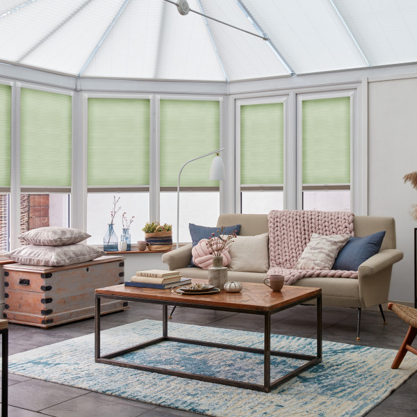 Pale green pleated cellular blinds, lowered over tall bay windows, filtering soft daylight; conservatory living room with beige sofa draped by a chunky pink throw, wooden coffee table and rug.