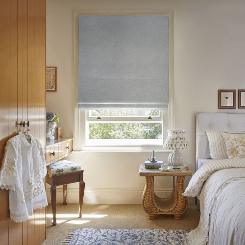 Grey textured Roman blind lowered over the window, partially open at the bottom filtering daylight. Cozy sunlit bedroom with wicker bedside table and lamp, bed, wooden wardrobe, leafy outside view.