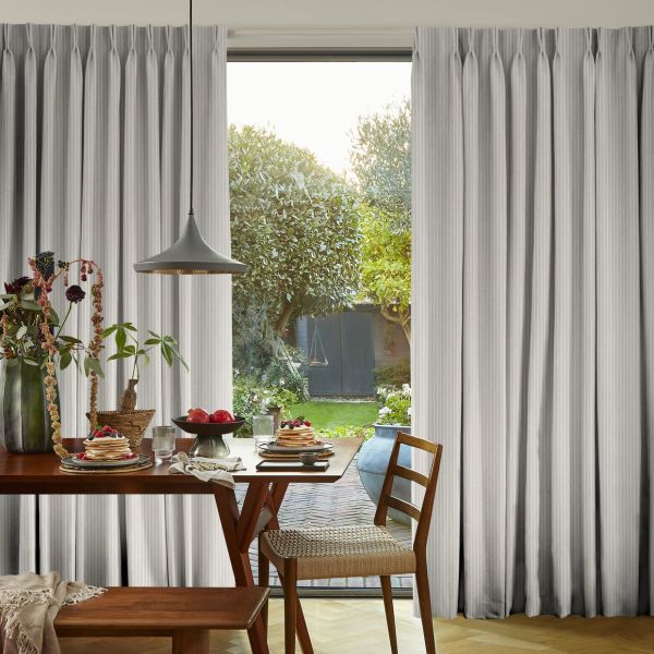 Light grey pinched-pleat curtains, floor-to-ceiling and parted centrally, reveal a glass sliding door; sunlight spills into a dining nook with wooden table, pendant lamp, wicker chair and garden beyond.