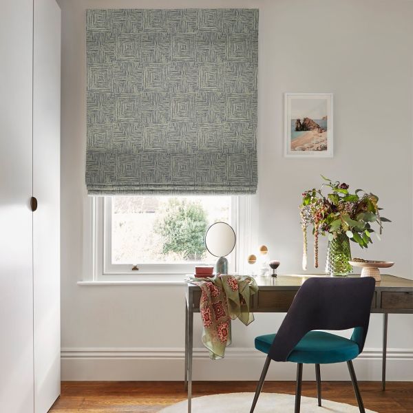 Roman blind with blue-gray textured square pattern, lowered and partially filtering daylight — a study window above a desk with mirror, chair, vase of flowers and framed beach print.