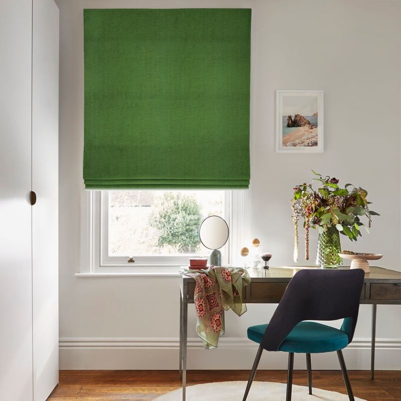 Green Roman blind — solid fabric with soft horizontal folds — is lowered, filtering daylight; a neat desk and teal chair sit beneath in a bright, minimalist home office with flowers.