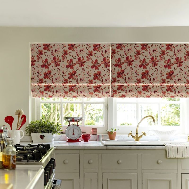 Pair of red-floral Roman blinds, folded and mostly lowered across two adjacent windows, filtering bright daylight; above a white kitchen sink and marble countertop with brass faucet, potted plants, stove.