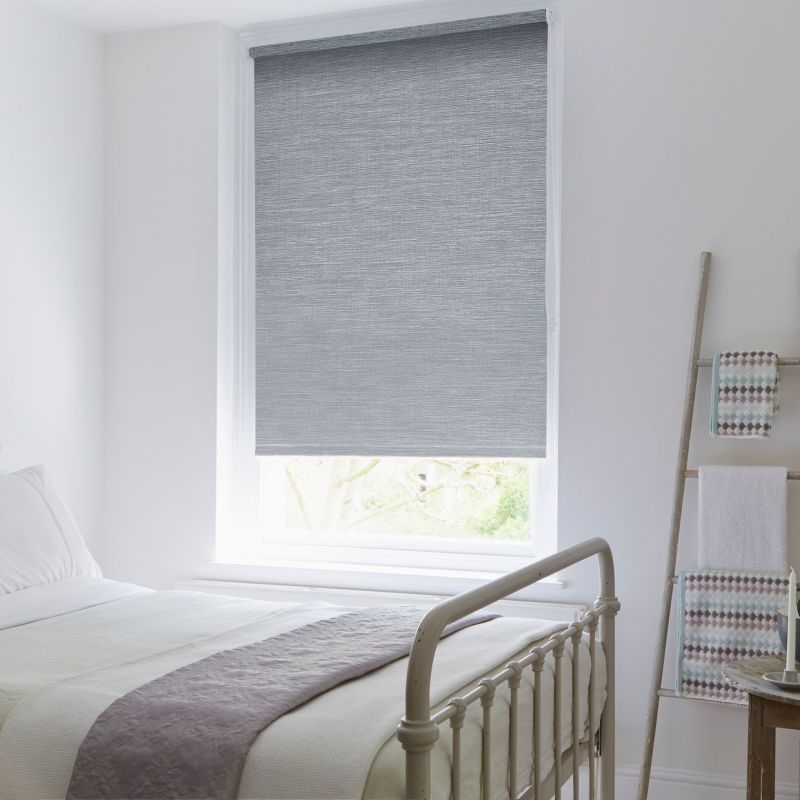 Grey textured roller blind lowered over a single sash window, filtering daylight; in a bright white bedroom with a metal bed frame, soft bedding, and a towel-ladder against the wall.