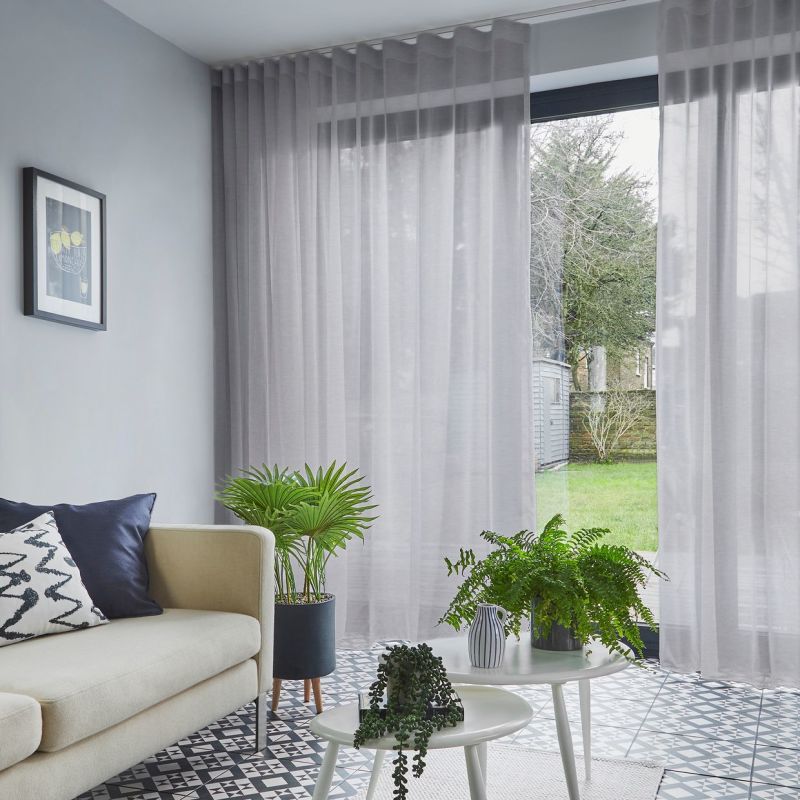 Sheer grey floor-to-ceiling curtains on a hidden track, filtering daylight and partially drawn to reveal a lawn and shed; living room with beige sofa, patterned tile floor and potted plants.