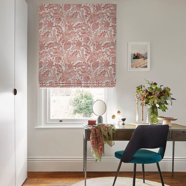 Roman fabric blind with pink floral-leaf pattern, lowered and partly folded, filtering soft daylight; positioned above a desk with teal chair, vase of foliage, and neutral walls in an office.