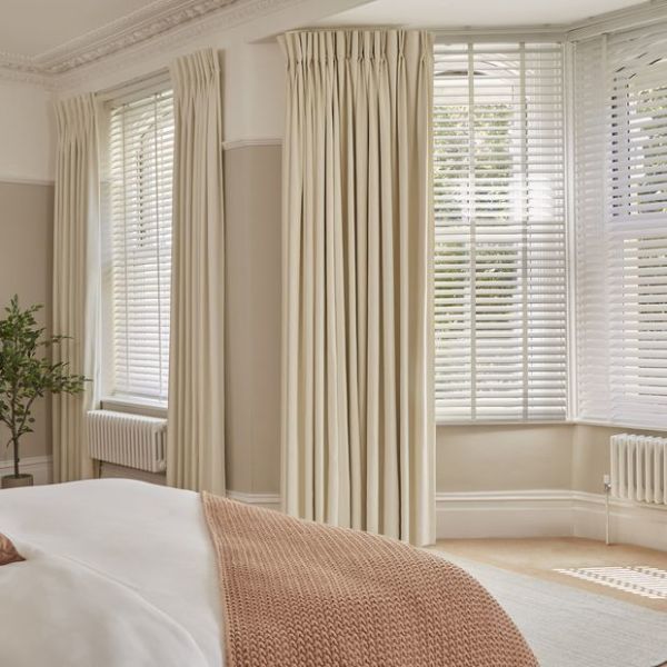 Cream pleated, floor-length curtains with white horizontal blinds covering bay windows; curtains pulled aside, blinds partially closed filtering soft daylight into a neutral-toned bedroom with bed and plant.