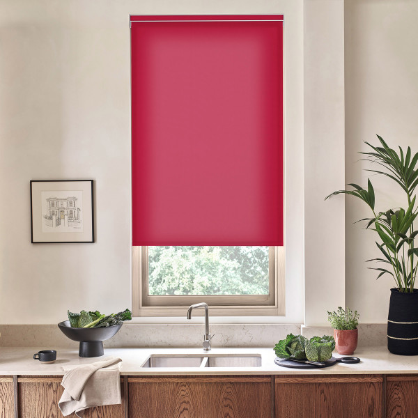 A red roller blind covers most of a kitchen window above a stainless faucet, with a countertop holding sink, vegetables in bowls, a towel, potted plants and a framed sketch.