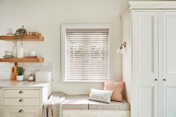 Horizontal pale wooden Venetian blinds, partially lowered and filtering soft daylight; a cozy kitchen nook with cushioned bench, pillows, floating wood shelves and white cabinetry. Text: "THE PIG", "BREAD".