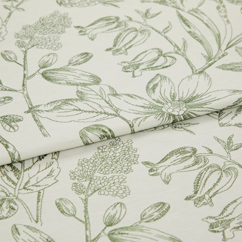 Folded curtain fabric with green line-drawn botanical print; overlapping fold reveals large floral and bud motifs, laid flat in a close-up studio view with soft, neutral lighting.