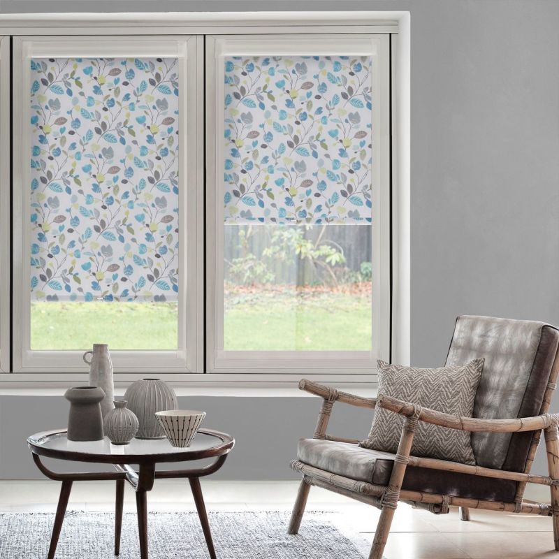 Floral leaf-pattern roller blinds covering twin windows—one fully lowered, one partially—filtering daylight; bright living room with gray walls, rattan armchair, round wooden table with ceramics, backyard lawn visible.