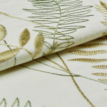 Cream linen fabric with gold and green embroidered fern and leaf motifs, folded to reveal raised satin and outline stitches catching light; close-up studio view with soft, diffuse lighting.