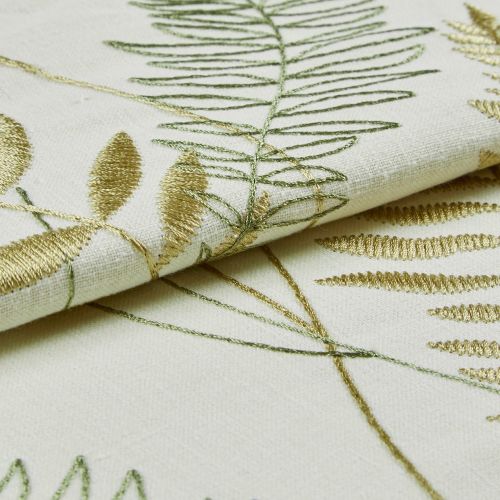 Cream linen fabric with gold and green embroidered fern and leaf motifs, folded to reveal raised satin and outline stitches catching light; close-up studio view with soft, diffuse lighting.