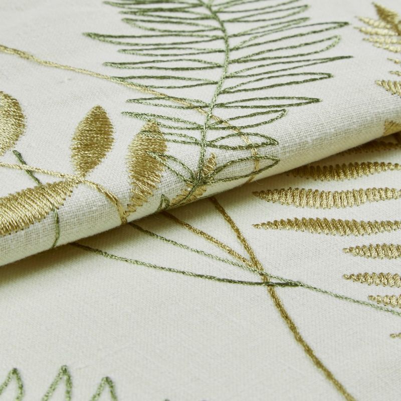 Cream linen fabric with gold and green embroidered fern and leaf motifs, folded to reveal raised satin and outline stitches catching light; close-up studio view with soft, diffuse lighting.