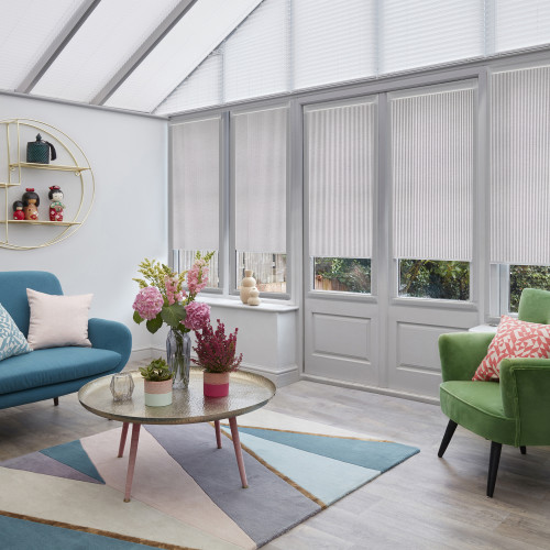 White pleated cellular blinds lowered across tall conservatory windows and door, filtering soft daylight; living room with teal sofa, green armchair, floral centerpiece, geometric rug, garden view.