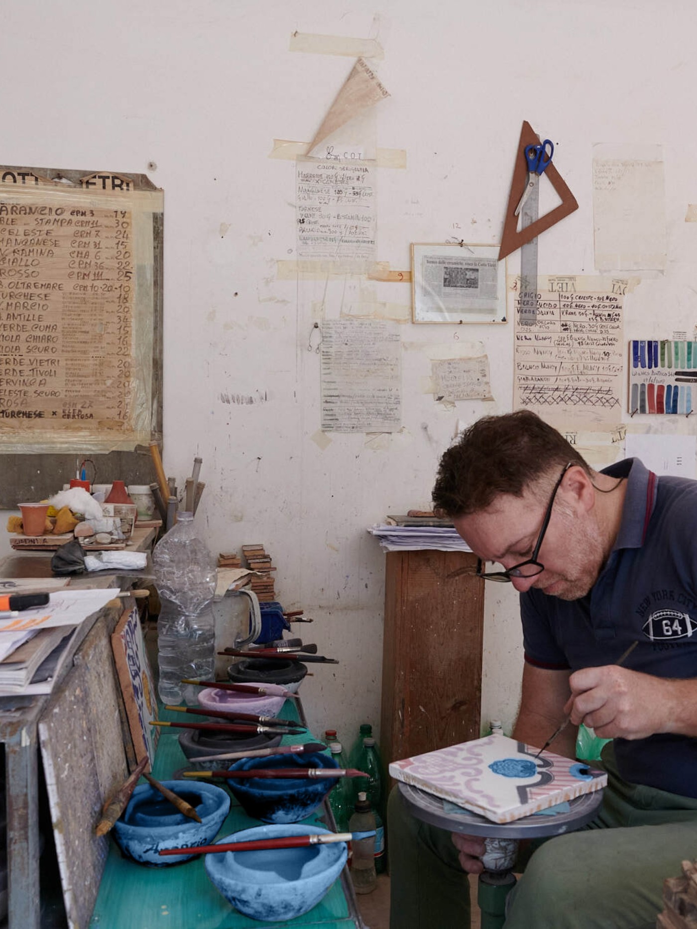 a man working on painting tile in a cluttered art studio.