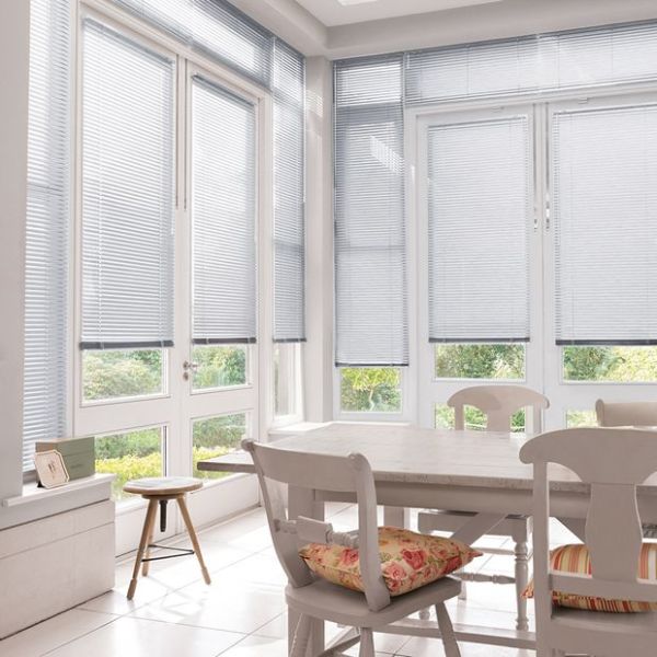 Light-gray horizontal Venetian blinds, fitted inside tall bay windows, partially lowered with closed slats filtering daylight; bright white dining nook with wooden table and chairs, garden visible beyond.