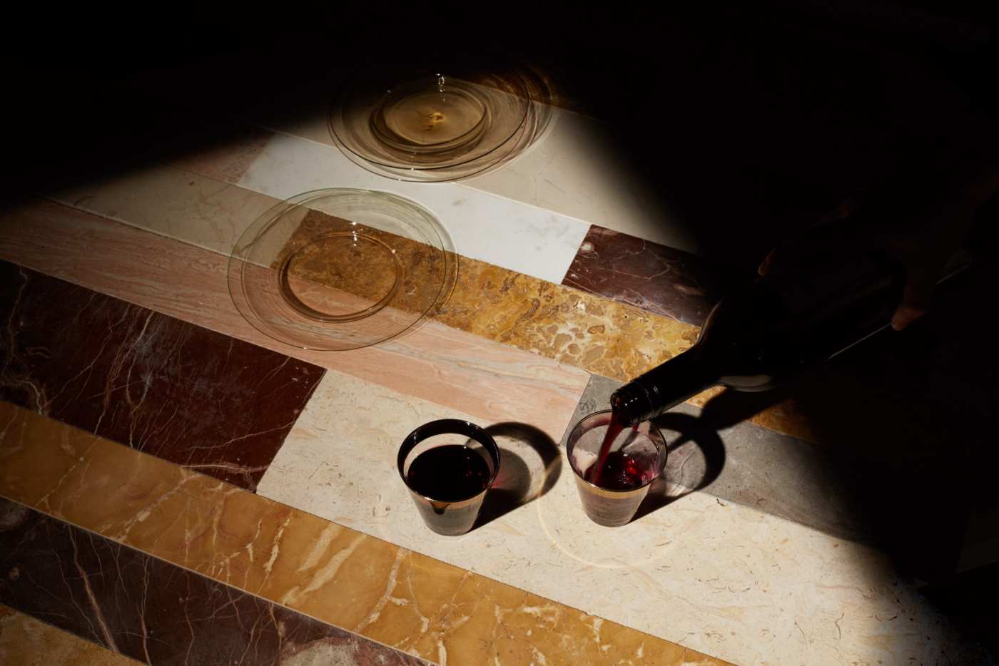 a person pouring wine into 2 glasses sitting on a tiled surface.