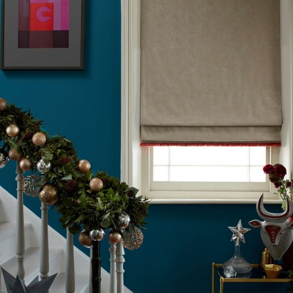Beige Roman blind with subtle texture and red fringe, lowered over a white-framed window, softly diffusing daylight in a teal-walled hallway beside a garland-wrapped staircase and metallic holiday ornaments.