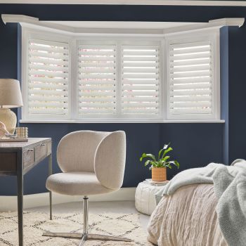 White plantation shutters covering a bay window, full-height panels with horizontal louvers — closed with slats partially open, filtering daylight into a navy-walled bedroom/study with beige chair, desk and bed.