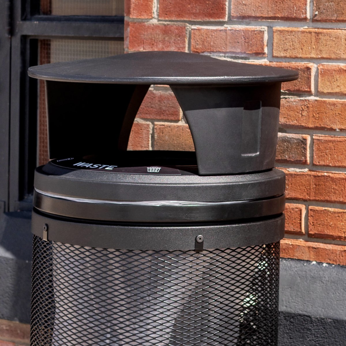 black metal and plastic waste bin with a canopy lid outdoors in front of a red brick wall
