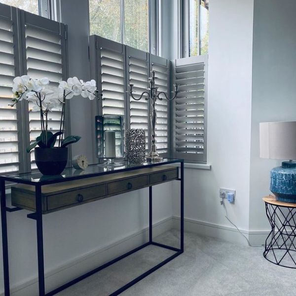 White plantation shutters covering lower window panels, angled louvers partially open, filtering soft daylight; in front, a slim metal-framed console table with orchid and decor, pale-carpeted modern sitting area.