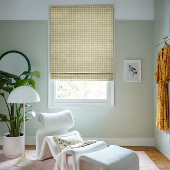 Beige woven Roman blind lowered over a single window, lattice-pattern filtering soft daylight, in a pale-green sitting corner with a white lounge chair, potted plant and a standing floor lamp.