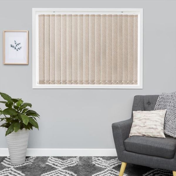 Beige patterned vertical blinds, scalloped bottoms, fitted inside a white frame — closed, softly filtering daylight. Context: grey-walled living room with armchair, cushion, potted plant and framed print.